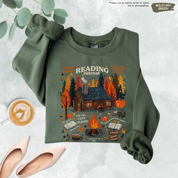 military green crewneck sweater with white Reading Retreat text above a graphic of a cabin in the woods. The trees are green, yellow, and orange and there are other objects around the exterior of the house, including two open books, a fire, a cup of coffee, pumpkins, and leaves