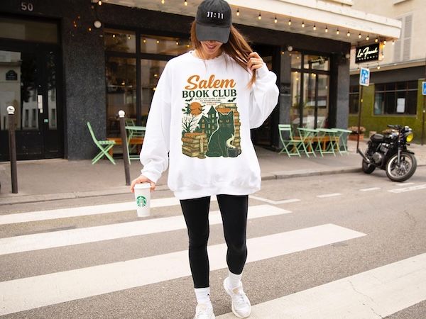 woman in a crosswalk wearing a white crewneck sweatshirt with Salem Book Club text above a graphic of a black cat, a stack of books, a Victorian house, and a witch on a broomstick