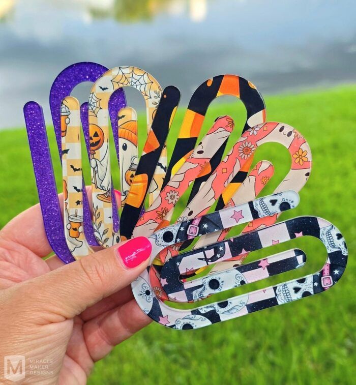 a hand holding five oversized plastic paperclip bookmarks with ghostly and halloween-themed prints