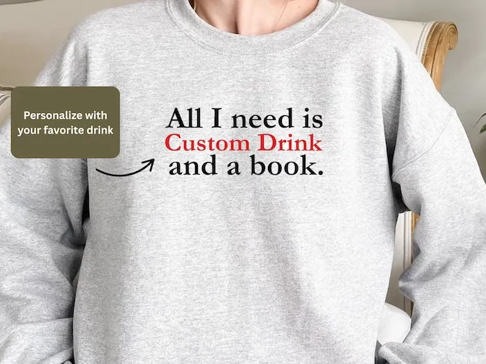 light heather grey crewneck sweatshirt with text that reads All I Need Is Custom Drink and a Book, where the Custom Drink text is bolded to indicate that text is customizable