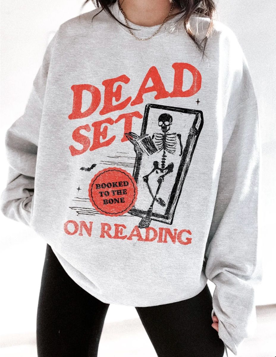 Image of a gray sweatshirt. It has a skeleton in a coffin and says "dead set on reading" in red. There's a red circle that says in black font "booked to the bone."