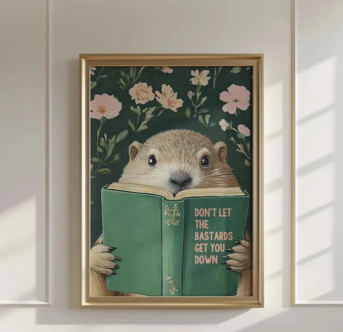 art print in a gold frame of a groundhog holding a green hardcover book with the title Don't Let the Bastards Get You Down