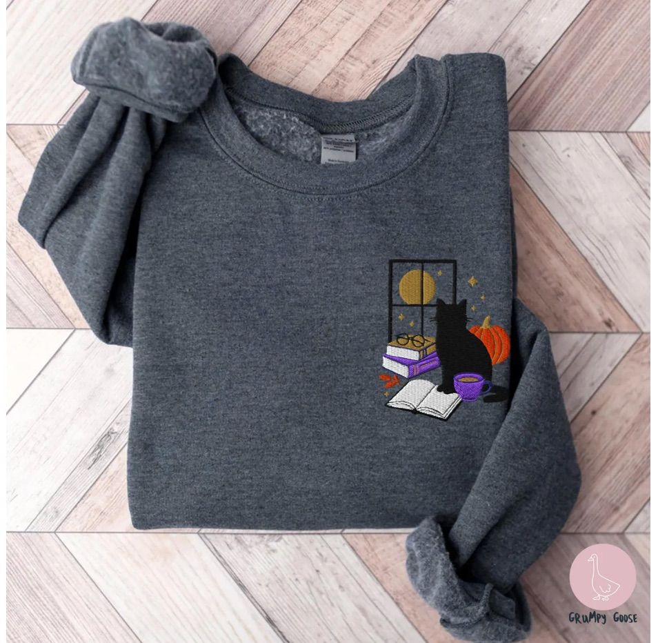 Image of a gray sweatshirt. There is a small embroidered cat with a pumpkin, coffee cup, and open book. 