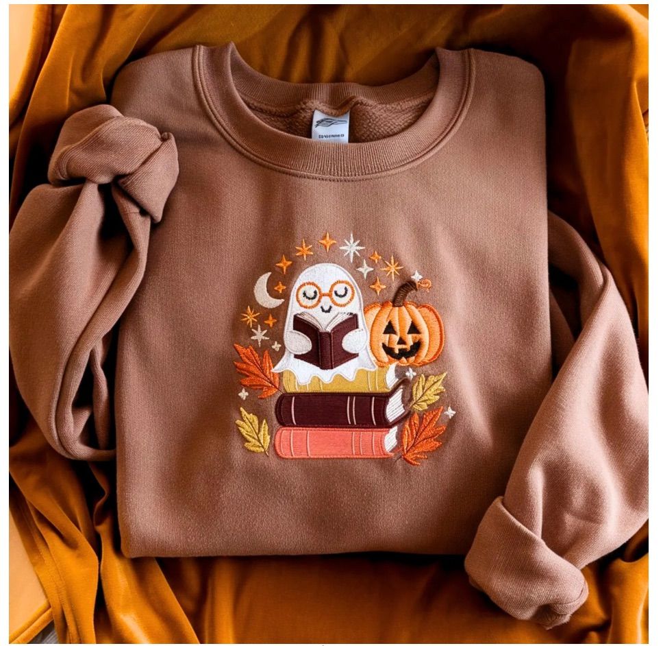 Image of a brown sweatshirt. There is an embroidered white ghost with orange glasses reading on top of a pile of books. 