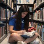 fair-skinned teen with long dark hair reading while sitting on the floor in a library