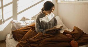 fair-skinned woman rading a book in bed with a brown comforter and pumpkins near her