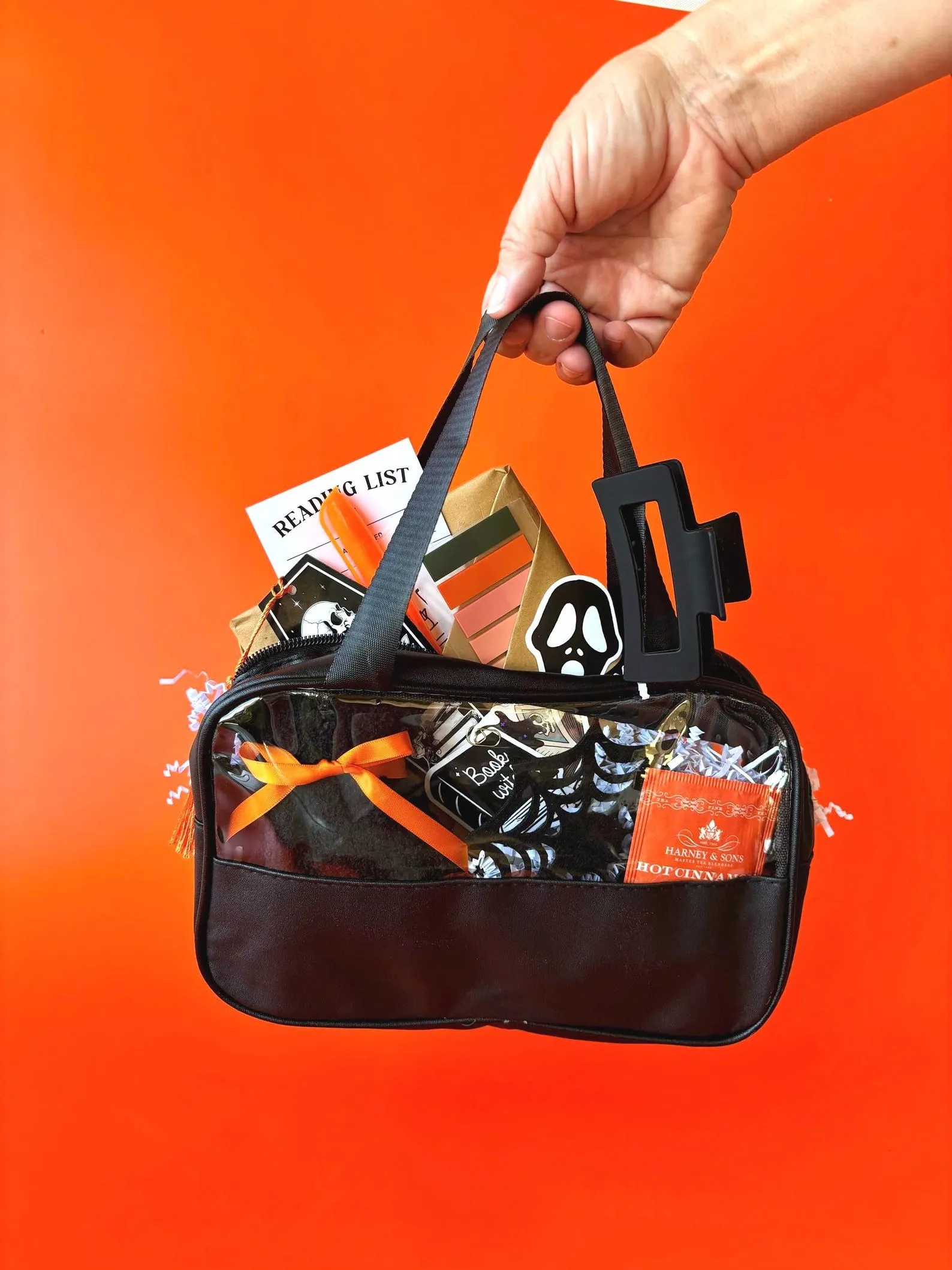 a hand holding a bag with halloween-themed goodies, including tea, snacks, and annotation supplies