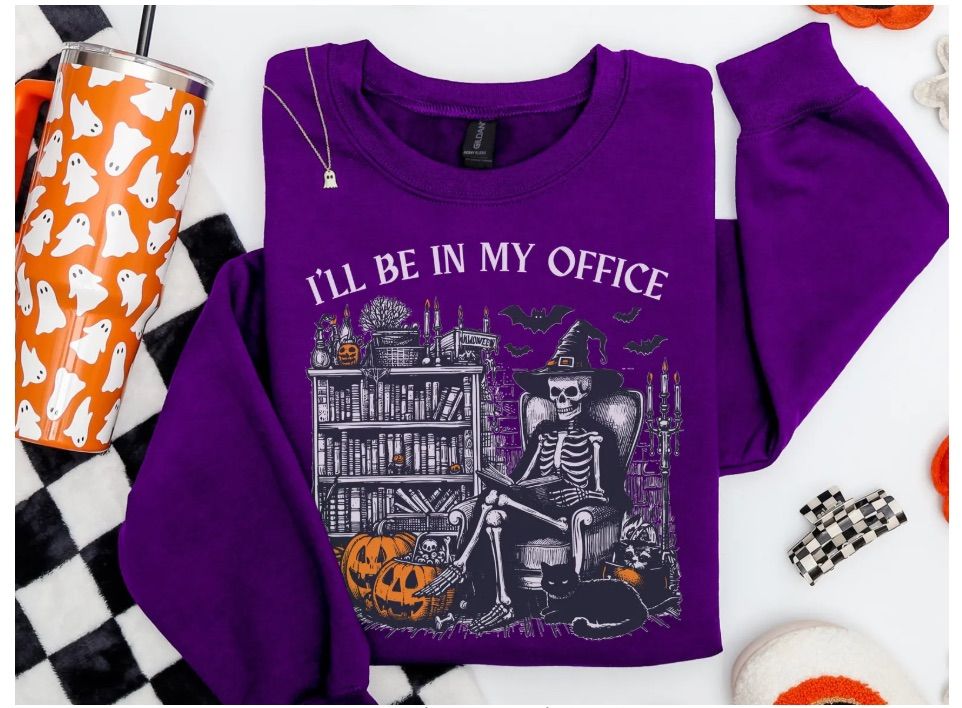 Image of a purple sweatshirt. It features a skeleton sitting in a library surrounded by pumpkins, bats, and a black cat. It says "I'll be in my office" in white font. 