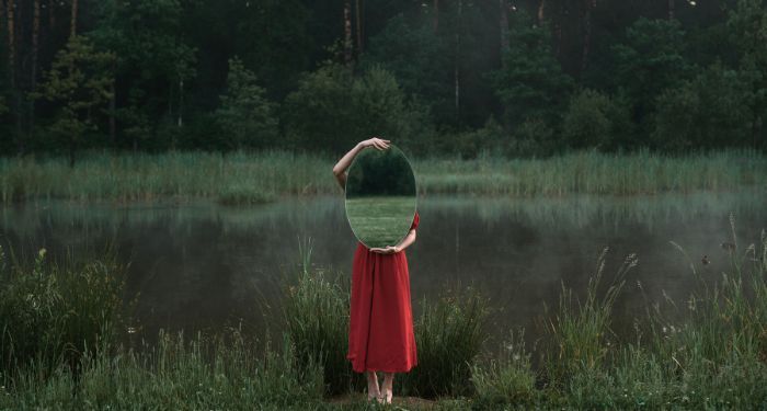 fair-skinned person in a red dress holds up an oval mirror in front of their face and torso while standing outside