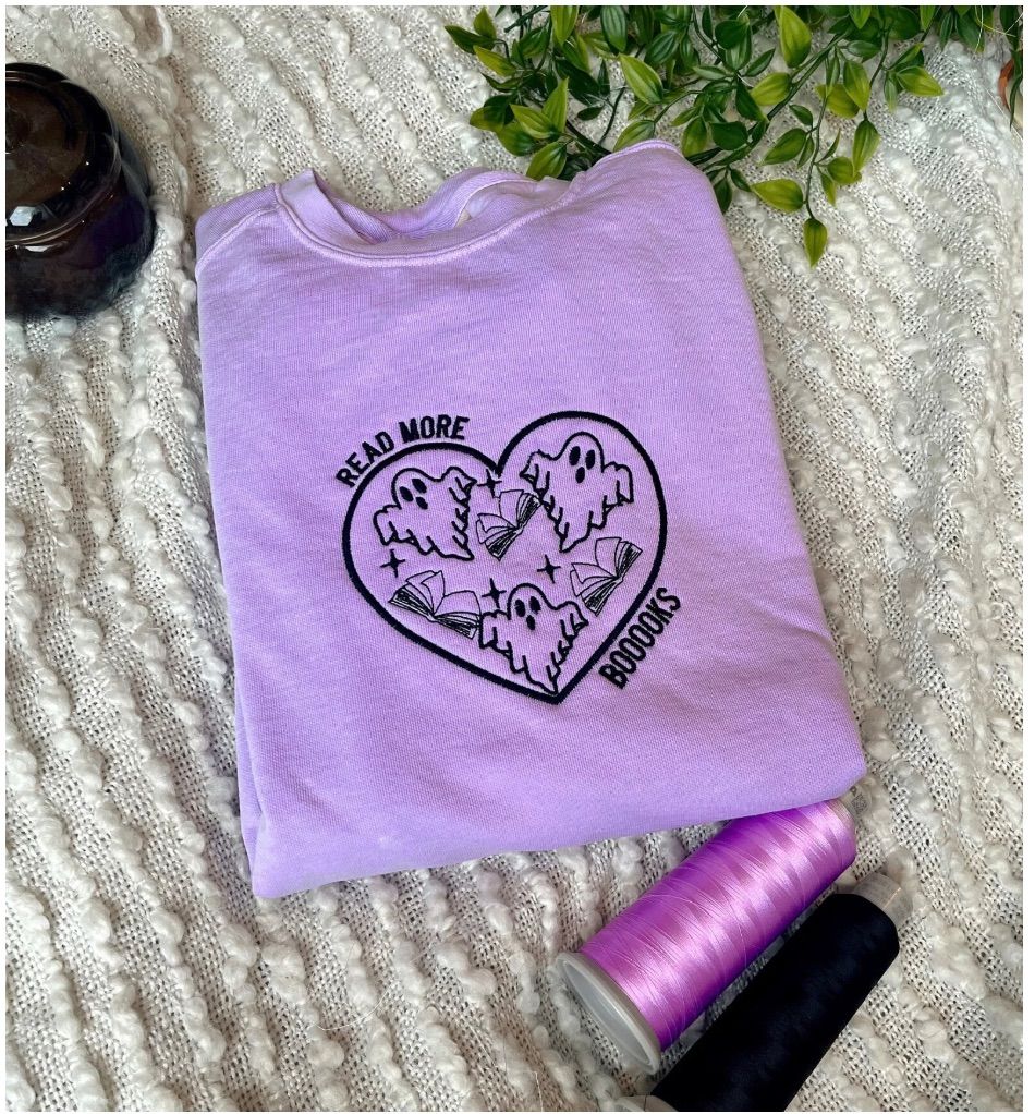 Image of a pale purple sweatshirt with black embroidery in the center. There is a heart with ghosts and books inside. Around the heart, it reads "read more boooooks."