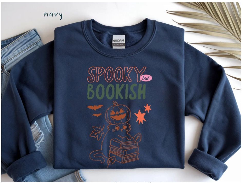 Image of a blue sweatshirt. It has a picture of a cat holding a stack of books. The cat has a pumpkin on its head. Above the image are the words "spooky but bookish."