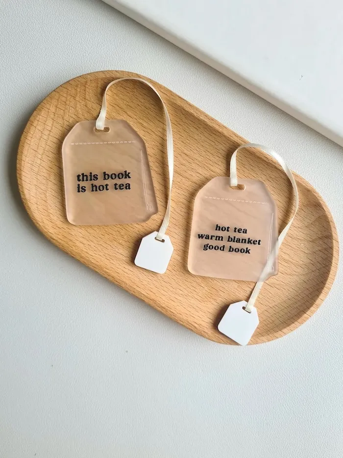 two clear acrylic bookmarks in the shape of tea bags resting on a wooden tray. both bookmarks have ribbons in them to look like strings on a tea bag and black text on them. One says This Book is Hot Tea, the other Hot Tea Warm Blanket Good Book