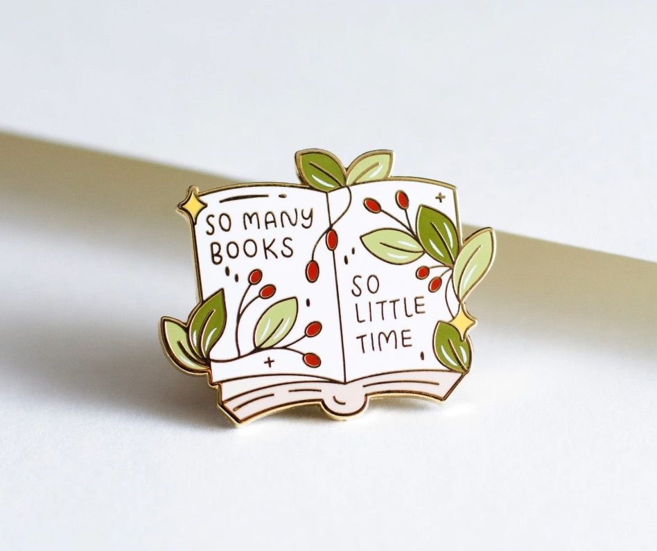 Image of an enamel pin. It looks like an open book with vines crawling on the pages, and it says "so many books so little time."