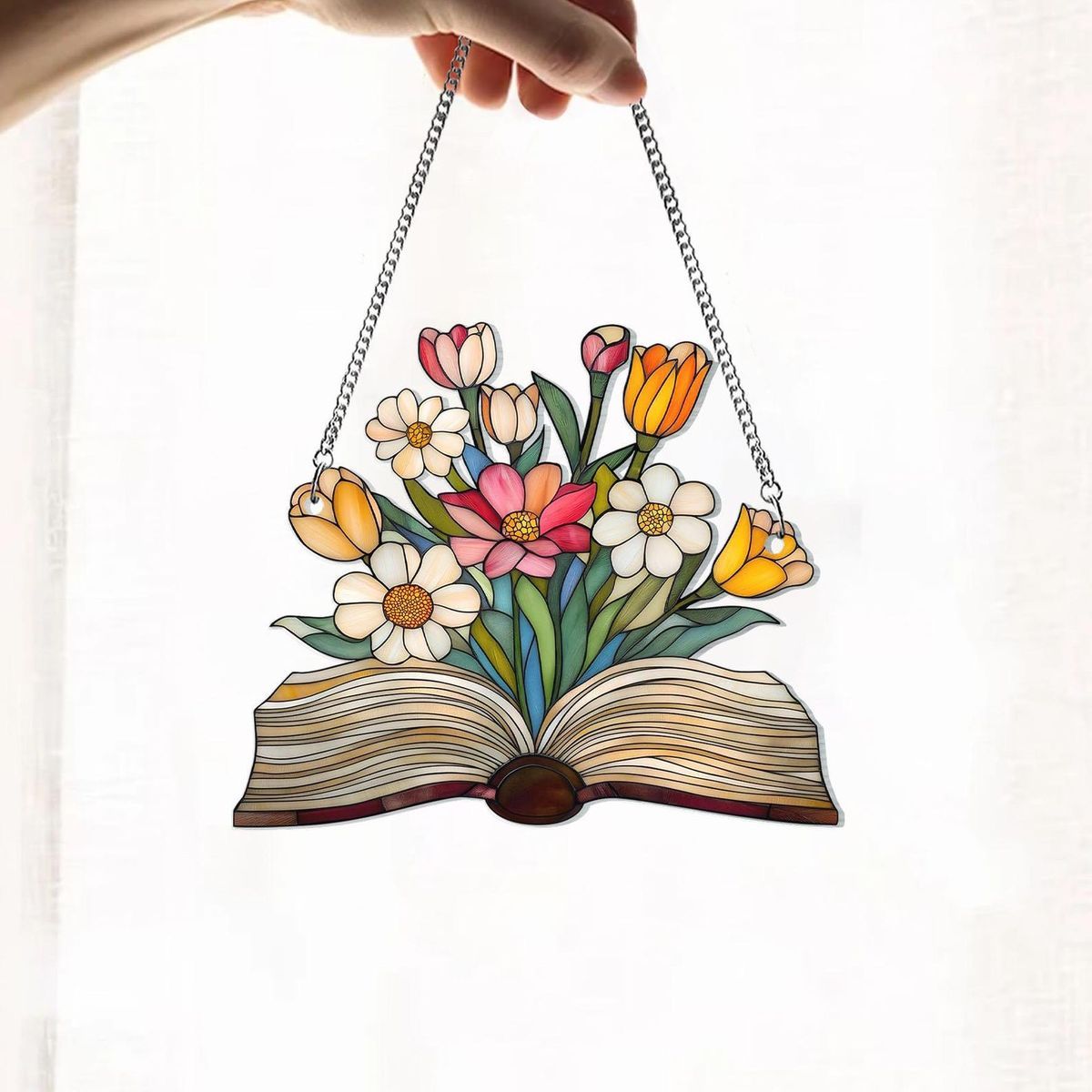 image of a hand holding a stained glass suncatcher. The suncatcher is shaped like an open book with a bouquet of spring flowers blooming from the pages and hangs from a metal chain