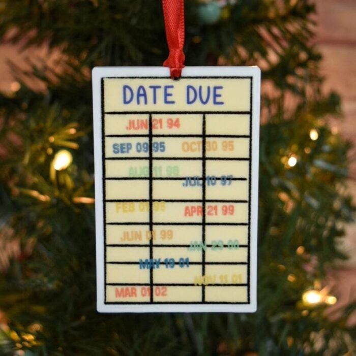 image of an ornament in front of a christmas tree. The ornament looks like a DATE DUE card with stamps and hangs from a red ribbon