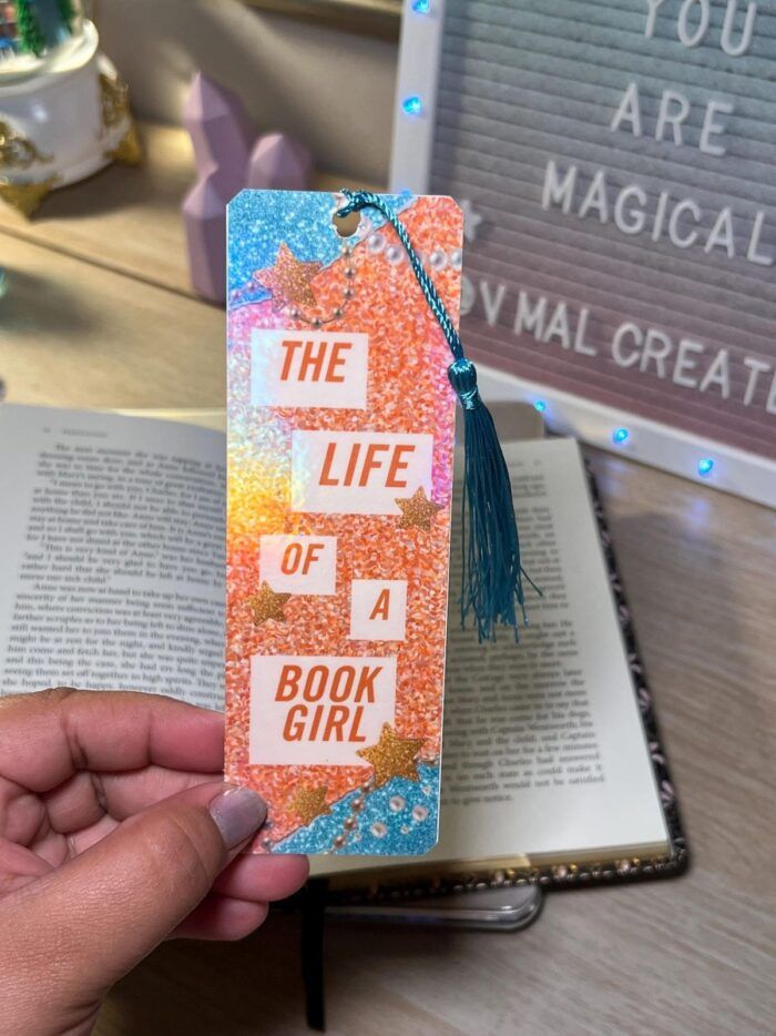 image of a hand holding a tassled bookmark that says "life of a book girl" with blue, orange, and pink decorations