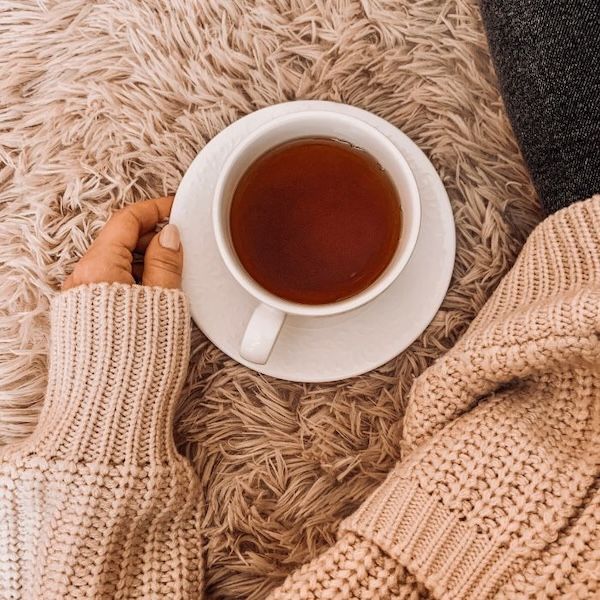 white teacup filled with dark amber-colored tea on a white saucer. the hand of a person in a chunky sweater is holding the saucer