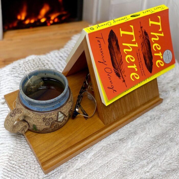 image of a wooden book stand near a fireplace. The book stand has a trianglar piece that holds a book open upside down, a mug holder, and a place for glasses.
