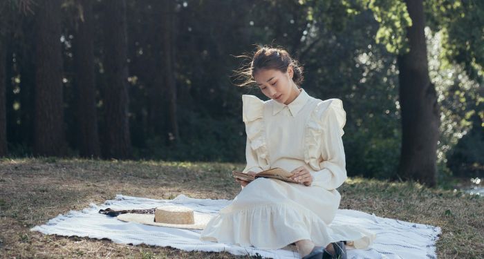 a fair-skinned Asian woman sitting on a blanket on the grass reading a book