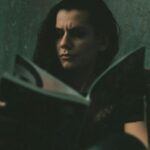 fair-skinned person with long hair reading a book against a greenish background